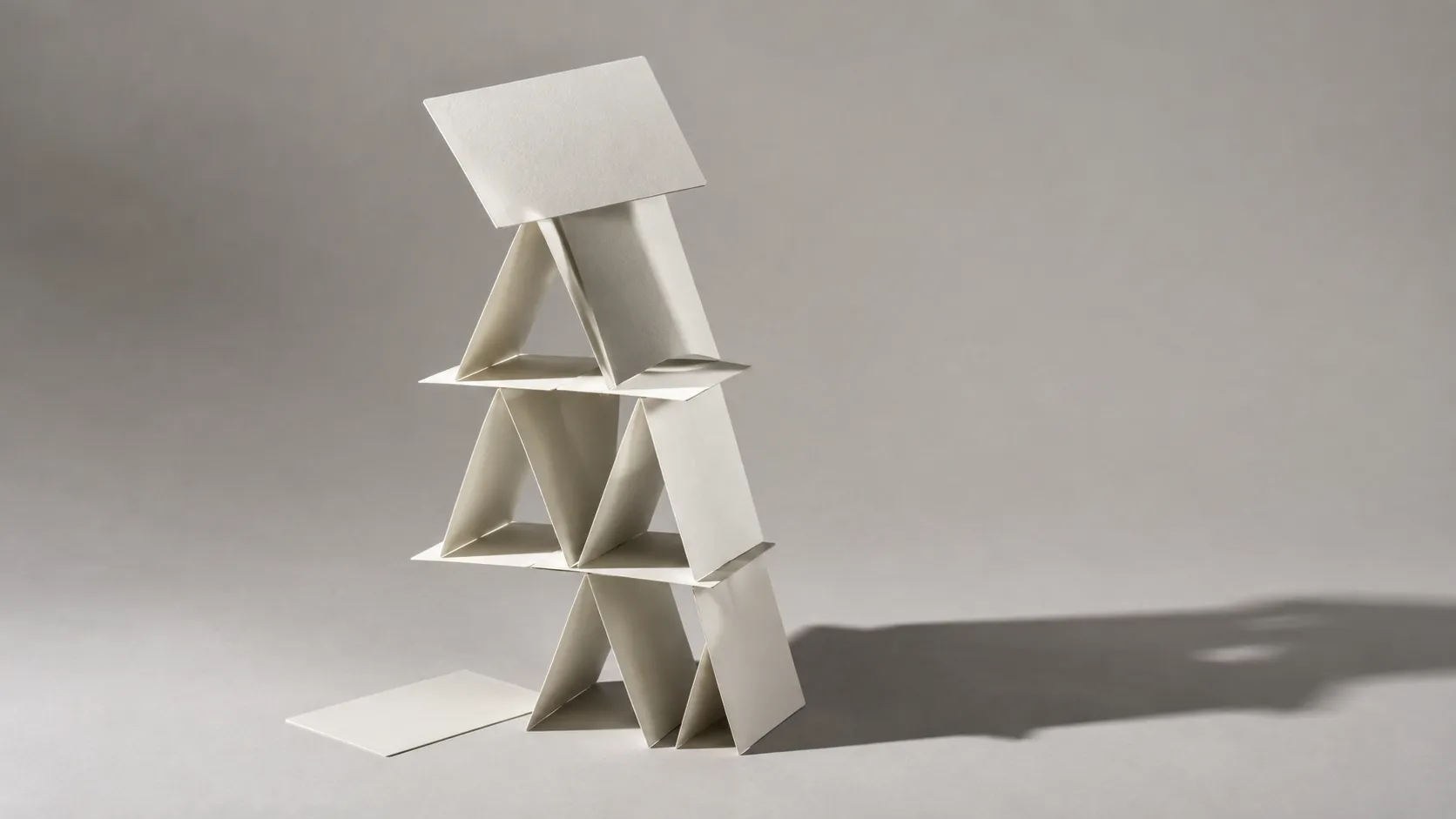 A house of cards built from plain paper sheets under hard studio light, with one sheet slipping out of the base
