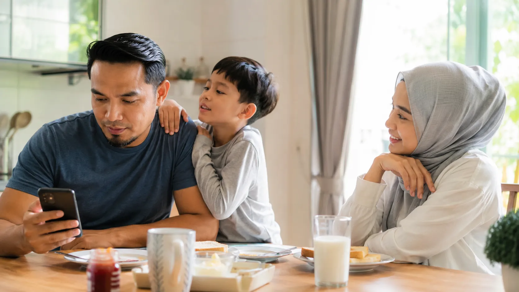 A bright family breakfast at home where a parent glances at their phone as their young child reaches for their attention