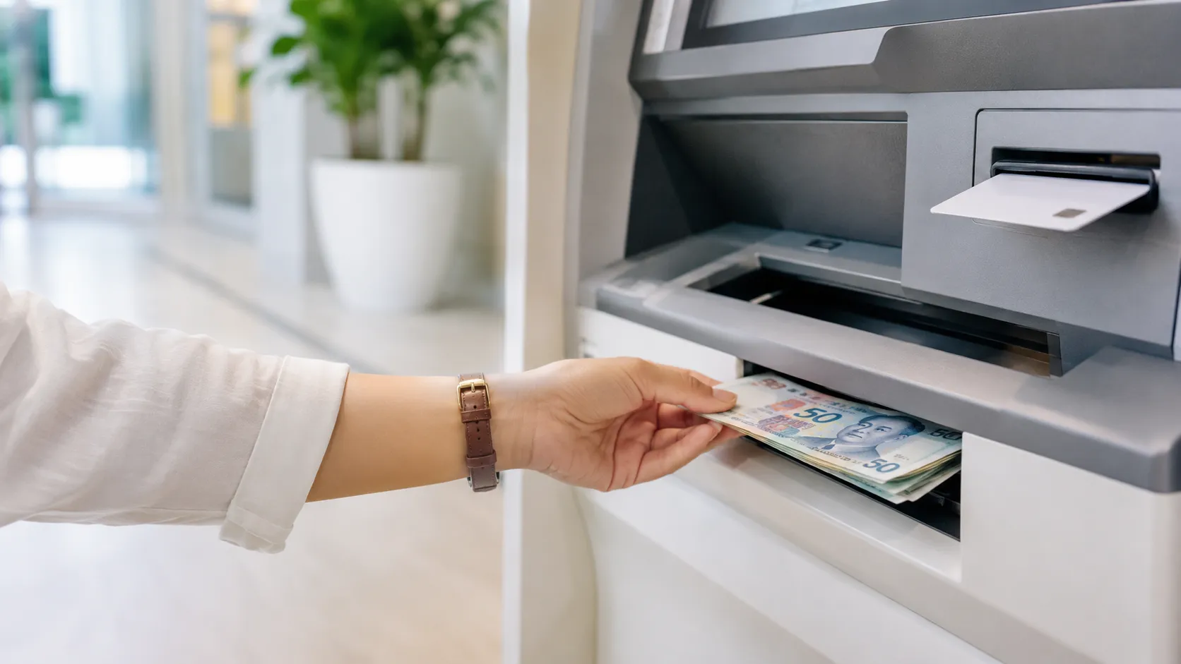 A traveller withdrawing Singapore dollar notes from a bright, modern ATM lobby