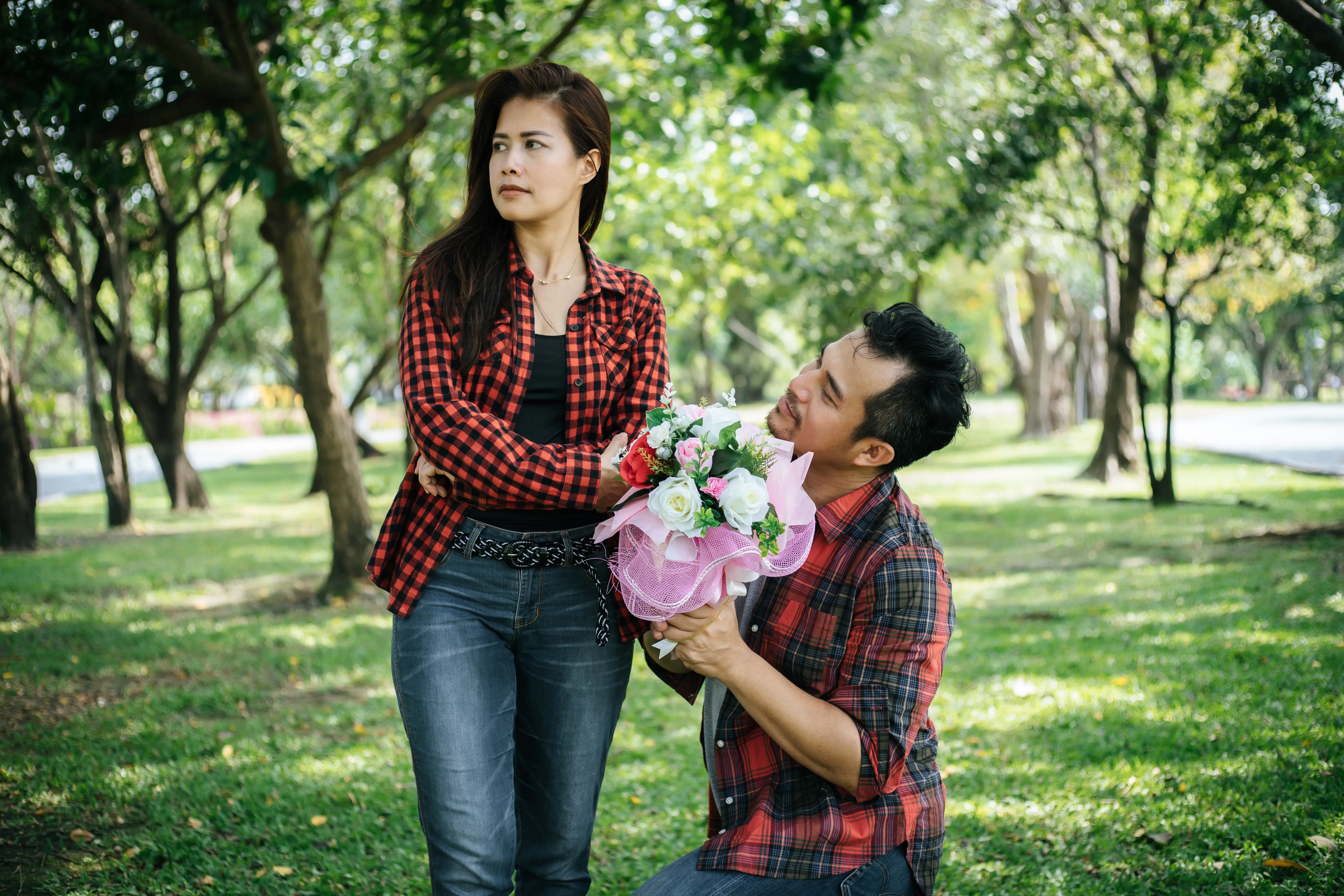 A man on his knees giving a bouquet of flowers to a woman with her arms crossed.