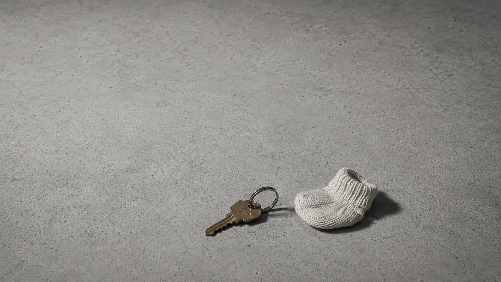 A single house key on a plain metal ring resting on a wide expanse of bare grey concrete, with one small knitted baby sock lying beside it